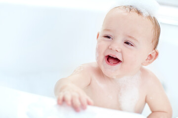 Bubbles, bathtub and baby laughing in bathroom for playing, washing and clean routine at home. Skincare, happy and cute boy toddler in water with foam for hygiene with growth and development in house