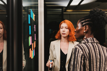 Two women collaborate in a modern office, discussing colorful sticky notes on a glass board, highlighting teamwork, planning, and creative brainstorming in a professional environment.