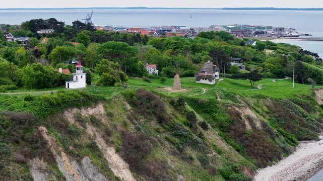Drone footage of lush green hills and steep coastal cliffs beside the sea, featuring small houses and a lighthouse near Ega and Mols Bjerge in Denmark.