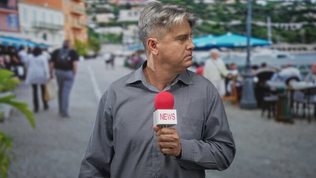 Man reporter holds microphone on a street promenade by a waterfront cafe during live news coverage, facing forward; serious duty.