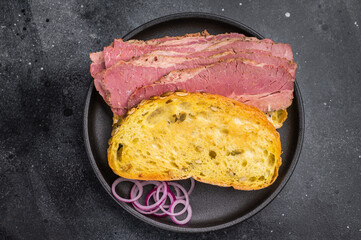 Smoked pastrami brisket slices in a hearty sandwich with seed crusted bread, onions and pickles on dark stone surface.