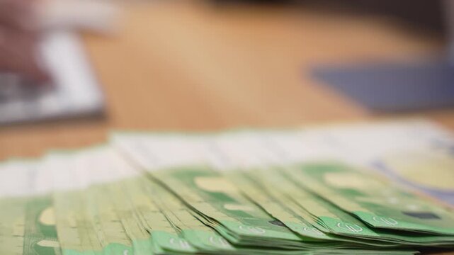 Fan of mixed euro banknotes lies on a wooden desk with a hand typing on a keyboard in the background. The camera pans from right to left across the colorful money.