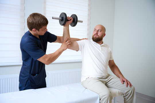 Physiotherapist assisting male patient with shoulder rehabilitation exercise using dumbbell. Man sitting on medical couch while performing physiotherapy workout under supervision of doctor.