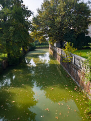 A beautiful old canal featuring a brick retaining wall and a classic stone balustrade along the bank surrounded by thick green trees and fallen autumn leaves