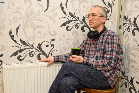 an adult man is sitting by a white radiator, crossing his legs, and holding a black mug - Powered by Adobe
