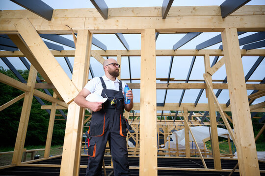 Construction worker taking a break and drinking water at wooden house frame construction site. Builder resting with hard hat and water bottle.