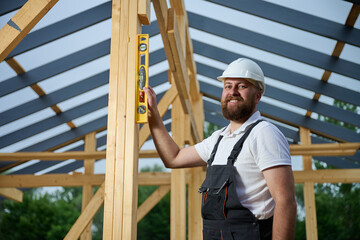 Construction worker using spirit level to check vertical alignment of wooden beam at frame house site. Builder wearing hard hat and safety glasses working on wooden structure outdoors.