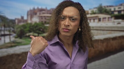 Hispanic transgender woman wearing purple shirt pointing finger at camera in old town street with stone wall; defiance.