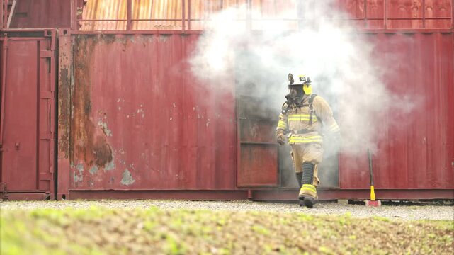 A firefighter in full protective gear runs out of a smoke-filled structure carrying an emergency axe. The scene captures urgency, determination, and active emergency response during a live training