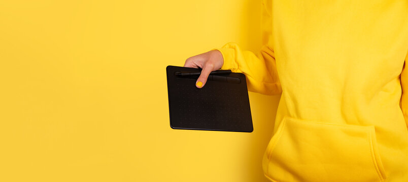 Close-up of a person's hand in yellow clothing holding a black graphic tablet against a yellow background, ideal for creative technology themes.
