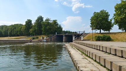 Fototapeta premium The river flows through a dam with a bridge and railings in the banks lined with concrete slabs with steps. Nearby there are grassy lawns with trees reflected in the water. Sunny summer weather