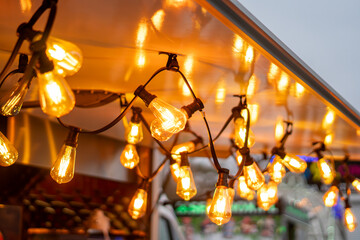 Warm decorative bulbs hanging beneath a Christmas market stall canopy, creating bright light reflections and a softly blurred festive background behind them