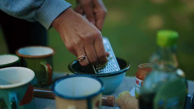 Close-up of a hand grating fresh ginger into a bowl outdoors with soft nature in the background.