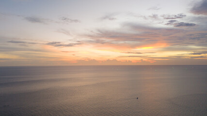Wide aerial view of a pastel sunrise or sunset over a calm ocean, highlighting a single small boat beneath a softly colored cloudscape.
