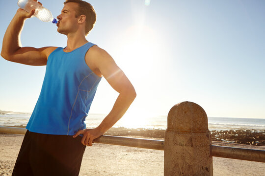 Fitness, break and man at beach drinking water, hydration and rest in training routine. Flare, low angle and athlete at ocean with sports bottle, h20 liquid and health detox in workout recovery.
