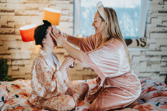 Two women in robes bonding as they apply face masks indoors during a relaxed and cheerful morning.
