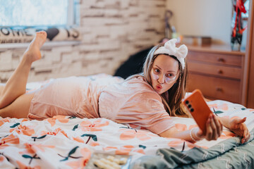 Woman lounging on bed, wearing skincare face patches and a headband, taking a selfie in relaxed and...