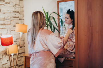 Two women wearing soft robes are sitting in front of a mirror discussing and smiling. The cozy indoor scene conveys feelings of relaxation, friendship, and comfort.