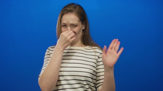 Woman in striped shirt pinches nose and winces to block pungent odor in bright blue studio setting; disgust.