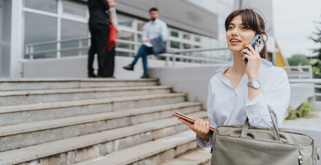 Confident businesswoman having a conversation on her phone while walking outdoors near offices, carrying a bag and documents. This portrays a professional work environment and modern office culture.