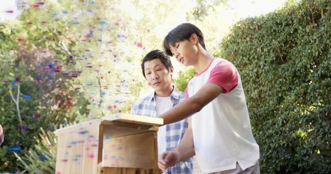 Father and son aligning wooden panel on box yard, following DIY plans, confetti drifting foreground
