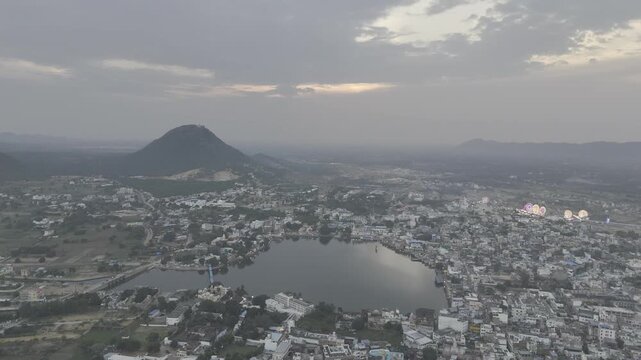 Aerial view of Pushkar Lake during the fair, showing cloud reflections on the water, glowing roller-coaster lights, lakeside houses and ghats, with the warm sunset over the Aravali hills in the back
