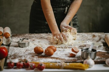 Close-up of baker's hands kneading fresh dough on a rustic wooden table, surrounded by baking ingredients like flour, eggs, and measuring cups, highlighting the homemade process of food preparation.