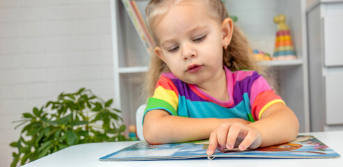 Adorable little girl in a bright t-shirt reading a book and pointing at the page.
