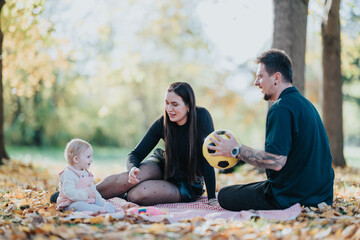 A young family sits on a checkered blanket in a sunlit autumn park. The baby watches as parents share smiles, while the father holds a soccer ball, creating a warm, playful outdoor moment.