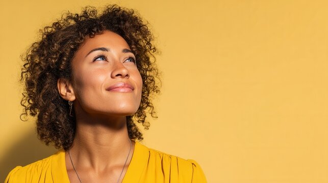 A cheerful woman with curly hair looks up with a smile against a bright yellow backdrop, conveying positivity and confidence.
