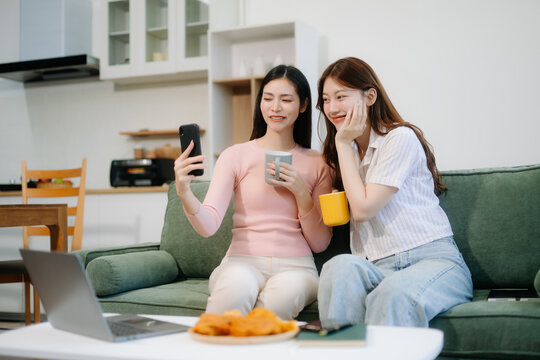 Two cheerful women sitting on a green couch, sharing ideas using a laptop and phone in a stylish home setup. - Powered by Adobe