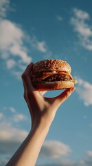 A hand raising a juicy hamburger with cheese and sauces against a bright, blue sky, symbolizing outdoor enjoyment.