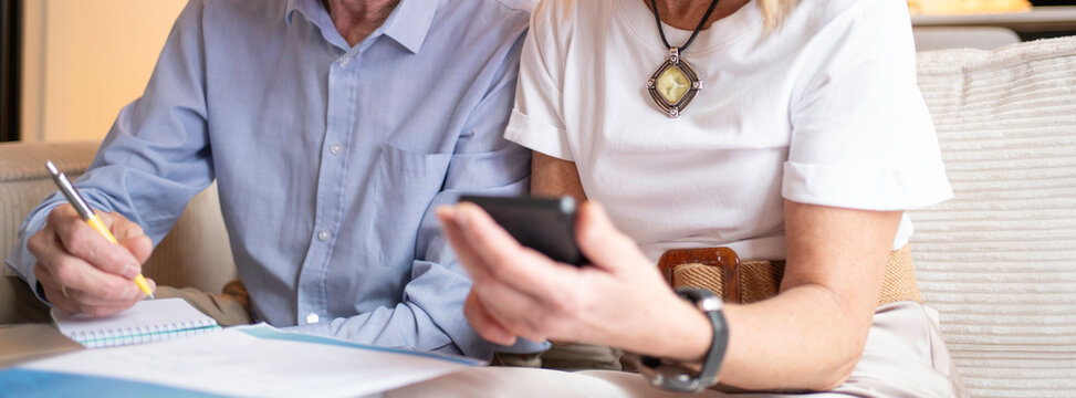 Close up of senior couple managing household finances, researching information on a smartphone, and writing notes