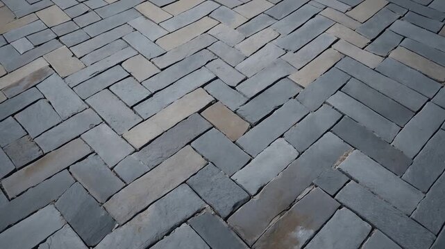 Close-up view of textured slate stone pavers arranged in a herringbone pattern showcasing cool gray and warm beige shades with natural light casting soft shadows across the surface emphasizing the