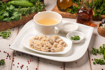 Pelmeni dumplings presented on plate alongside hot clear broth fresh herbs and creamy sauce