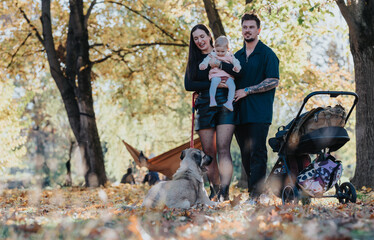 A family strolls through a sunlit autumn park. Parents hold their baby while a dog sits nearby, a stroller rests on colorful fallen leaves, capturing a warm, intimate moment of togetherness.