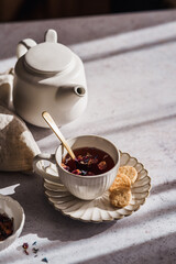 Eine Tasse Fr&uuml;chtetee mit Bl&uuml;ten und eine wei&szlig;e Teekanne im Morgenlicht auf einem grauen Tisch. Fr&uuml;hst&uuml;ck, Hei&szlig;getr&auml;nk.