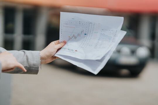 A close-up of hands holding and presenting financial data charts printed on paper. The scene is set outdoors, showcasing business communication and data analysis.