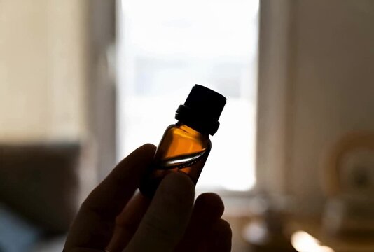 Amber Essential Oil Bottle Displayed in Silhouette with Hand Holding the Small Medicine Container in Front of Bright Window Light at Home 4k