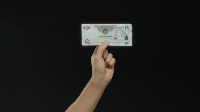 Hand holding a united arab emirates dirham banknote against a black background symbolizes currency and finance concepts.