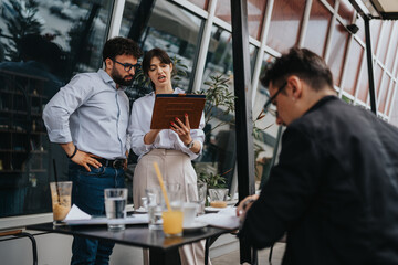 Two business associates, standing outdoors, confer over a tablet. The setting includes a casual meeting atmosphere, with seated and standing participants collaborating effectively.