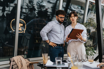 Two coworkers are seen discussing work using a tablet device in an outdoor cafe, surrounded by a modern environment and items such as drinks and documents, conveying collaboration and productivity.