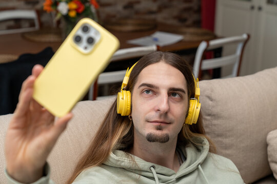 Young man with long hair relaxing on a comfortable couch, listening to music with headphones, taking a selfie