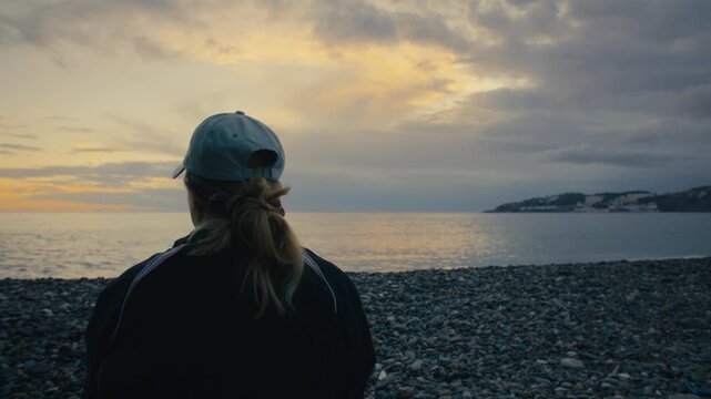 A woman sits facing the ocean during a dramatic sunset. Soft light, solitude, and quiet reflection on a rocky shoreline.