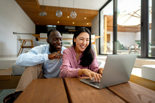 Happy young diverse, multiethnic couple planning budget, reading good news on laptop at home