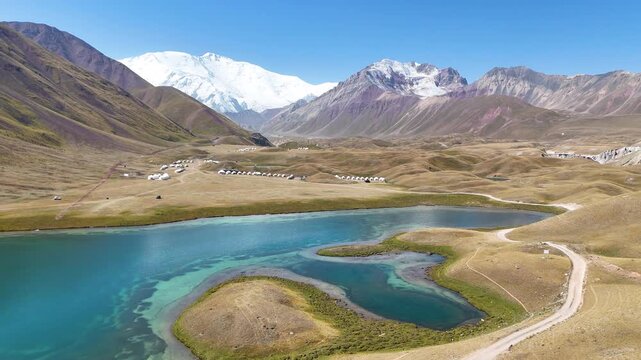 Beautiful view of Tulparkul Lake in a mountain valley with Lenin Peak and a snowy ridge in the background. A dirt road leads toward the shore where traditional Kyrgyz yurts stand peacefully.