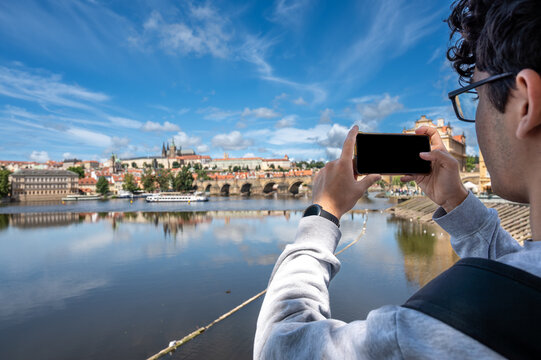 Prague, Czech Republic, August 9, 2023. A young Caucasian man takes photos with his cell phone of the enchanting view of the castle and cathedral dominating the hill. A lifestyle using technology.