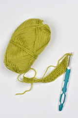 A ball of olive-green yarn, a crochet hook, and a small working swatch are displayed together on a white background