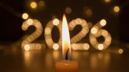 Warm candlelight from a single flame illuminates a dark room with the year 2026 shining in golden bokeh lights for a New Year celebration