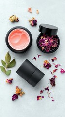 A top-down view of two black cosmetic jars, one filled with pink cream and the other with dried rose petals, surrounded by scattered dried rose petals and a gre